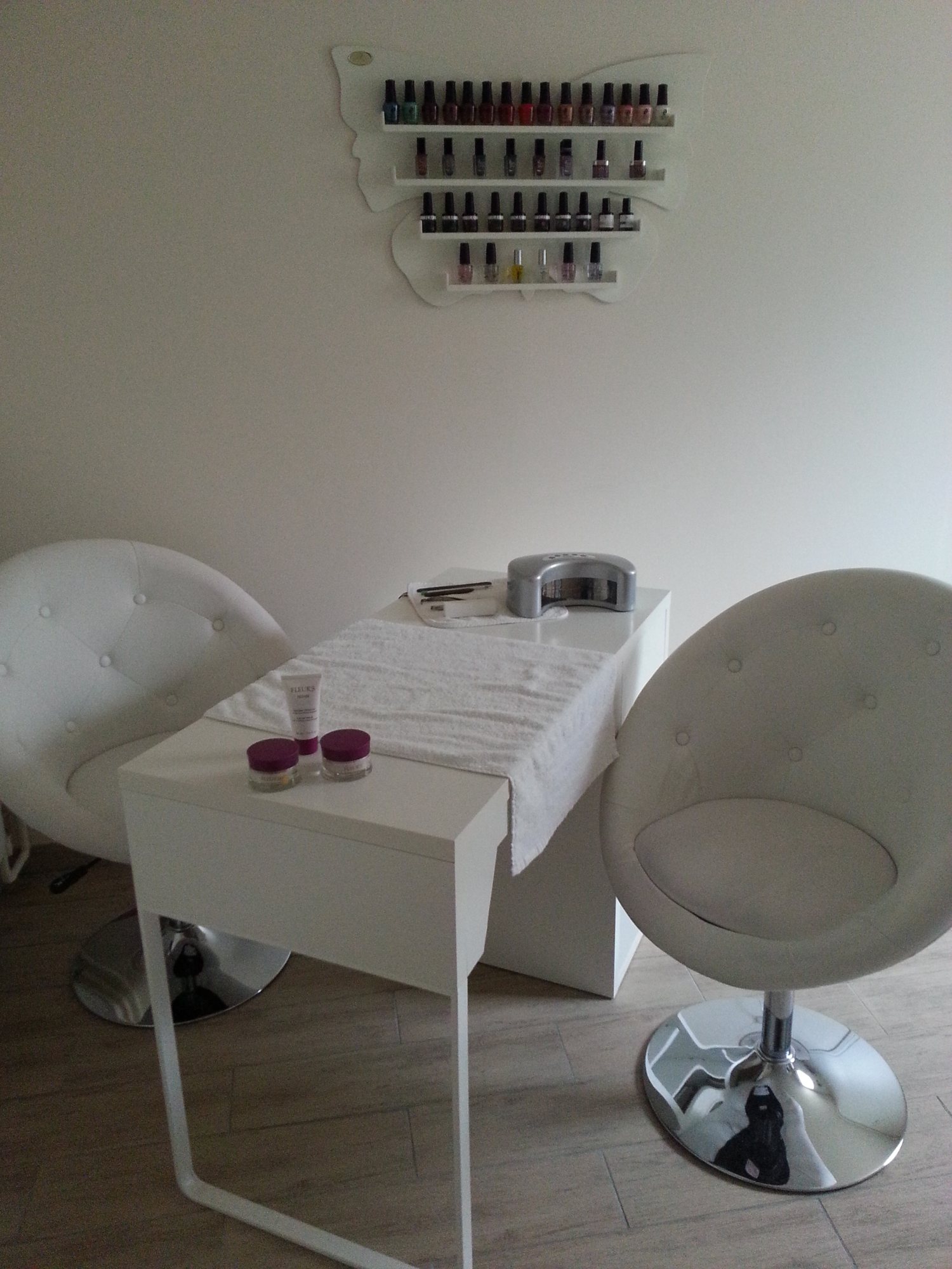 Un coin manucure épuré avec une table blanche, une lampe à ongles, deux fauteuils ronds blancs et un présentoir mural en forme de papillon garni de vernis.